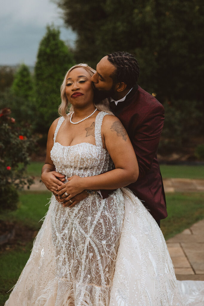 Groom hugging the bride from behind and kissing her cheek as she smiles with closed eyes, her beaded gown sparkling in the light.