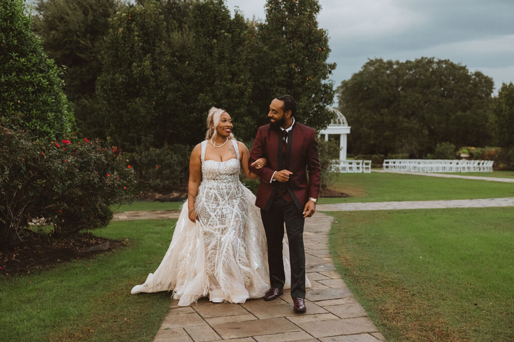 Couple walking hand in hand along a stone path, both smiling brightly, with wedding ceremony chairs and a gazebo in the distance.