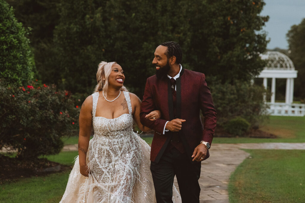 Bride and groom walking arm in arm along a garden path, smiling warmly at each other, with lush greenery and a white gazebo in the background.