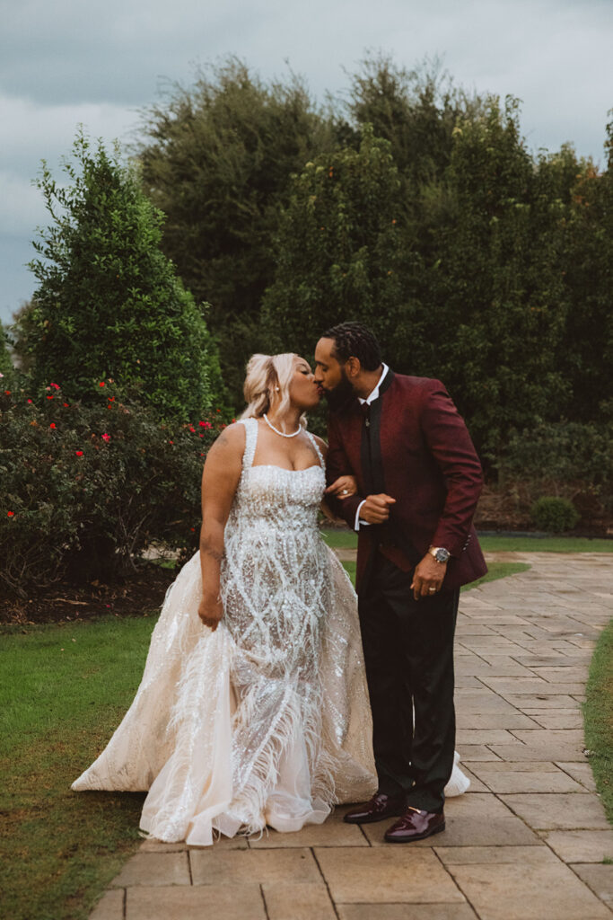Bride and groom sharing a kiss on a garden path, holding hands, with vibrant greenery and rose bushes on either side.