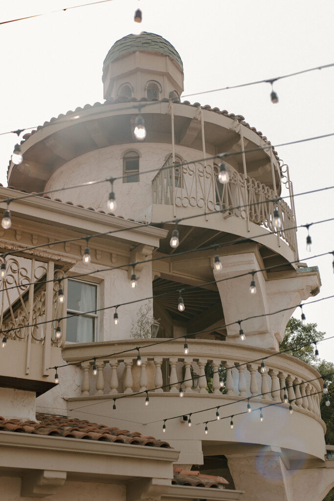 Mediterranean-style tower with a domed roof and wraparound balcony, viewed through strings of hanging café lights glowing softly in the evening light.  
