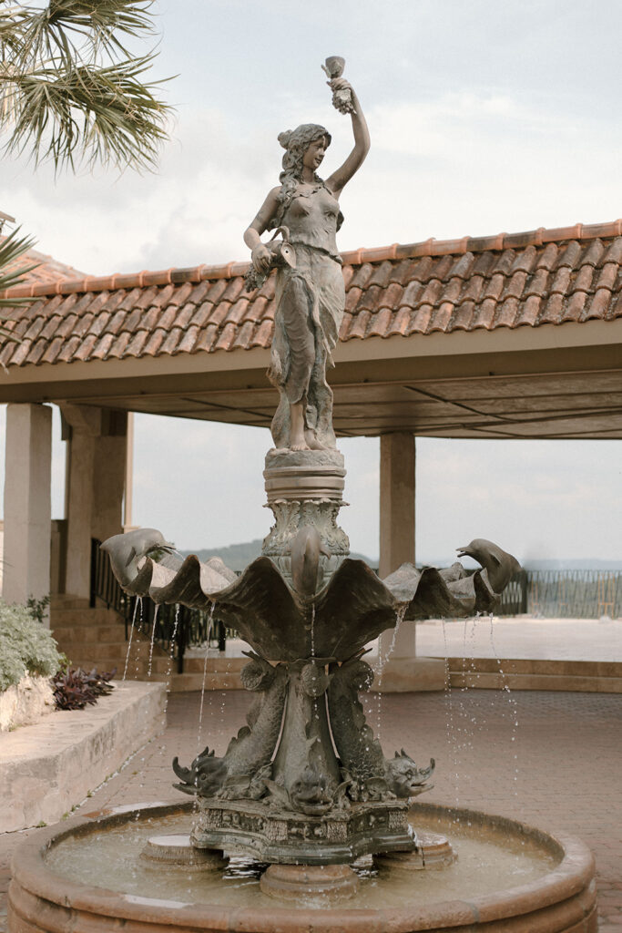 Outdoor bronze fountain statue of a woman raising a cup, with water flowing from sculpted dolphins into a round stone basin, set against a tiled-roof pavilion and hill country views.