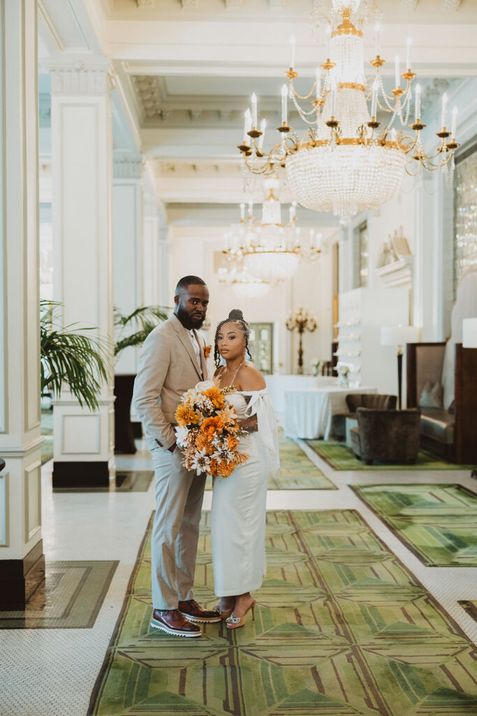 Bride and groom stand close together in an elegant hotel lobby, the bride holding a large bouquet of orange and white flowers beneath sparkling chandeliers.
