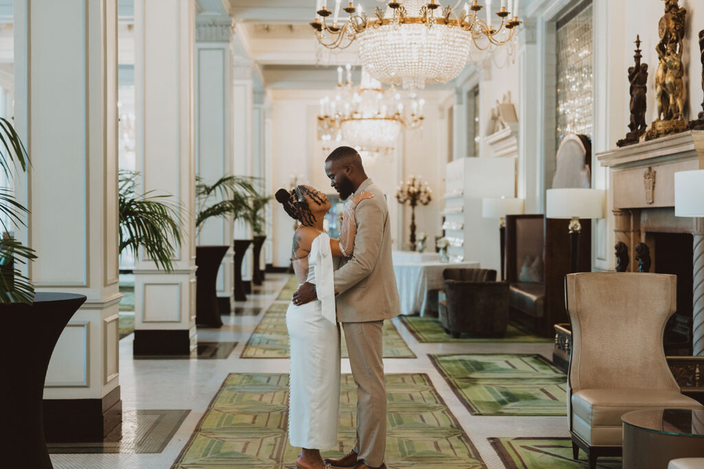 Bride and groom embrace in the center of a grand hotel lobby with chandeliers overhead, standing on patterned green carpet.