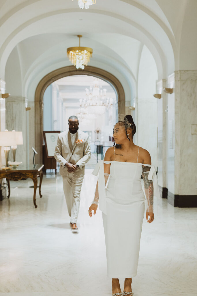  Bride in a modern white wedding dress and birdcage veil walks through an elegant marble hallway with chandeliers, as the groom in a tan suit follows behind.