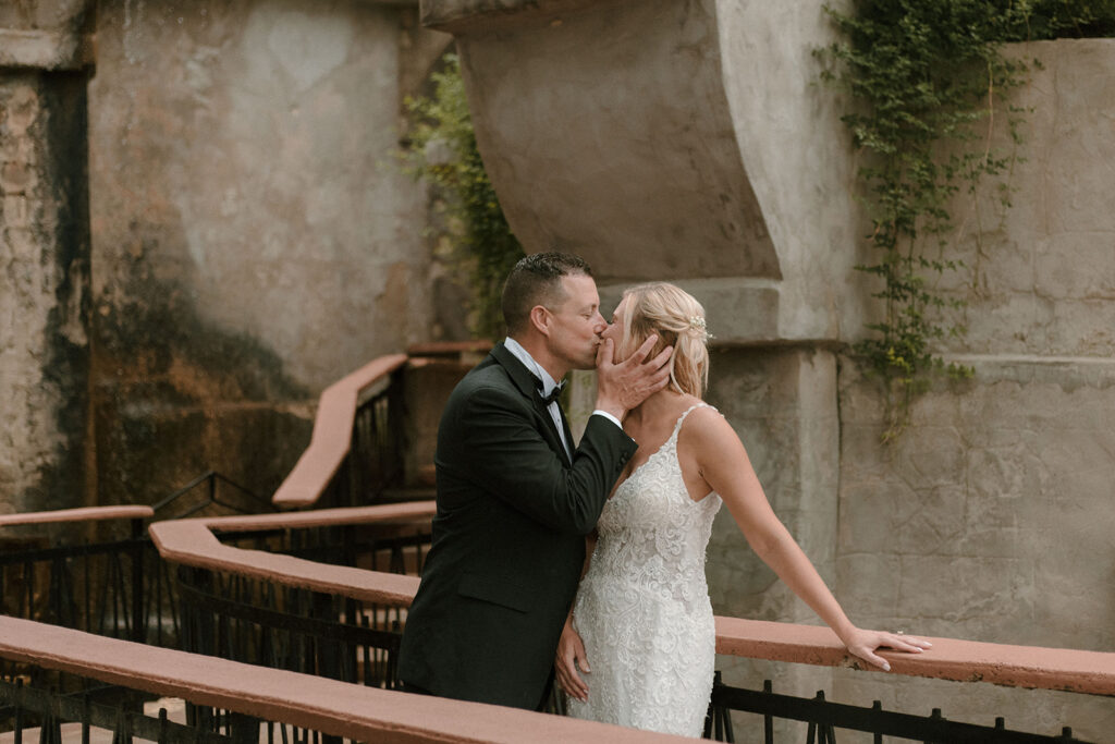 Groom in a black tuxedo gently holding the bride’s face as they share a kiss on a rustic stone balcony with terracotta railings and creeping vines.