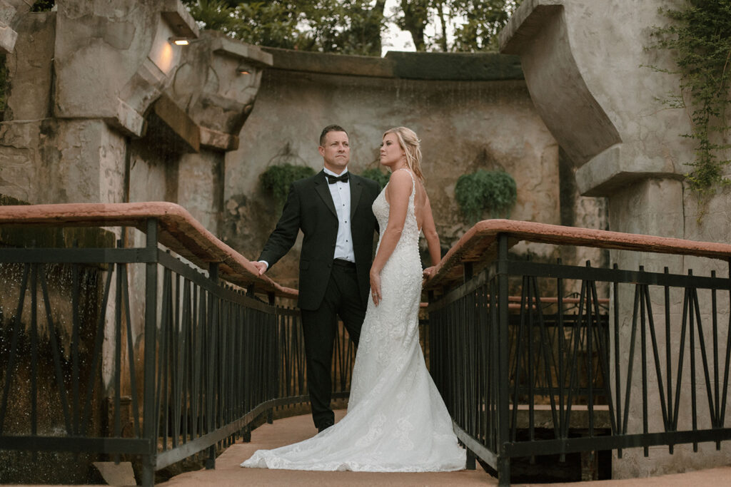 Bride and groom standing together on a narrow bridge with terracotta railings and black iron accents, framed by weathered stone walls and soft evening light.