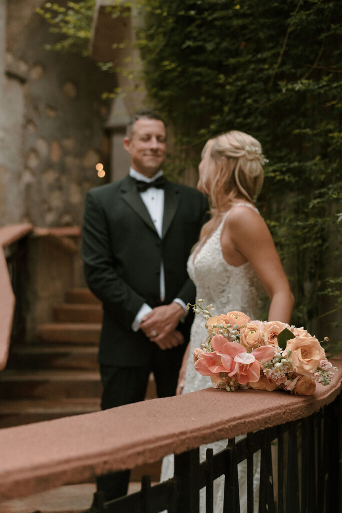 Peach and blush wedding bouquet resting on a terracotta railing in the foreground, with the bride and groom smiling at each other in the softly blurred background.