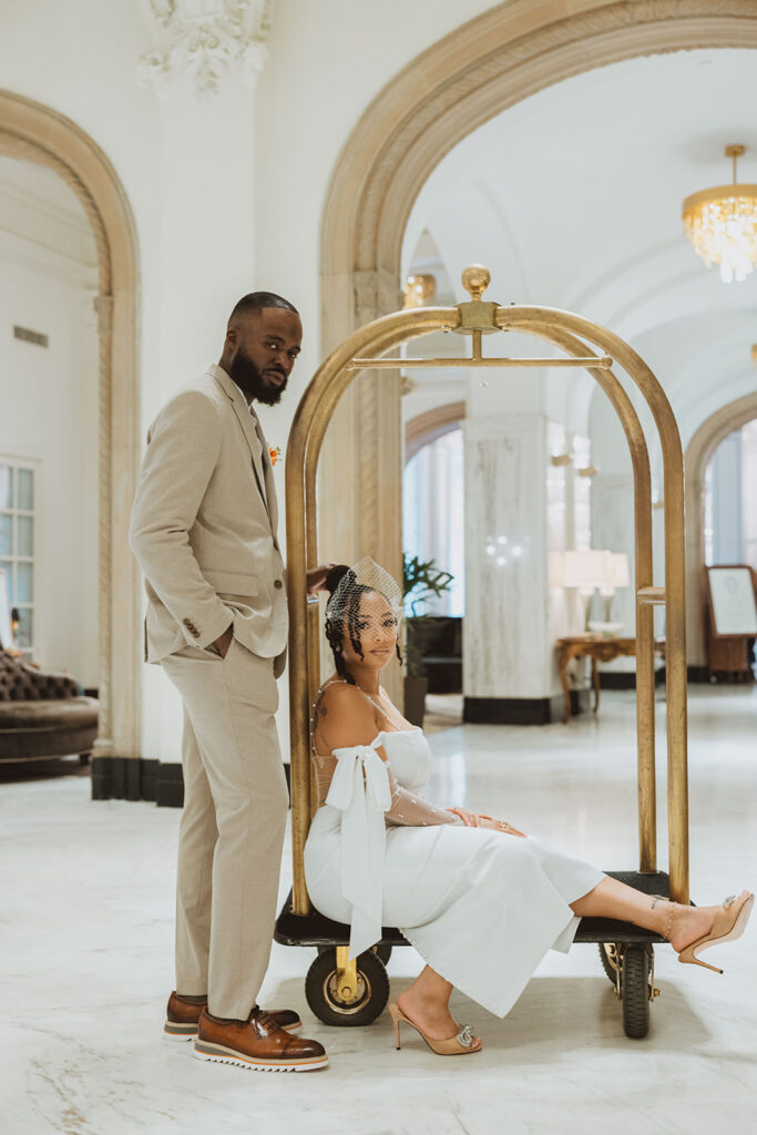 Bride and groom pose on a luggage cart in the archway of a luxury hotel with marble floors