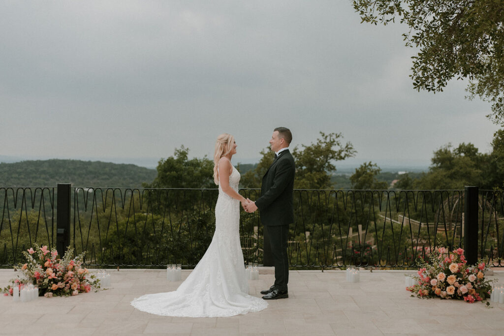 Bride and groom holding hands during their wedding ceremony on an outdoor terrace, with floral arrangements and candles framing the scene, overlooking a lush green hill country landscape under an overcast sky.