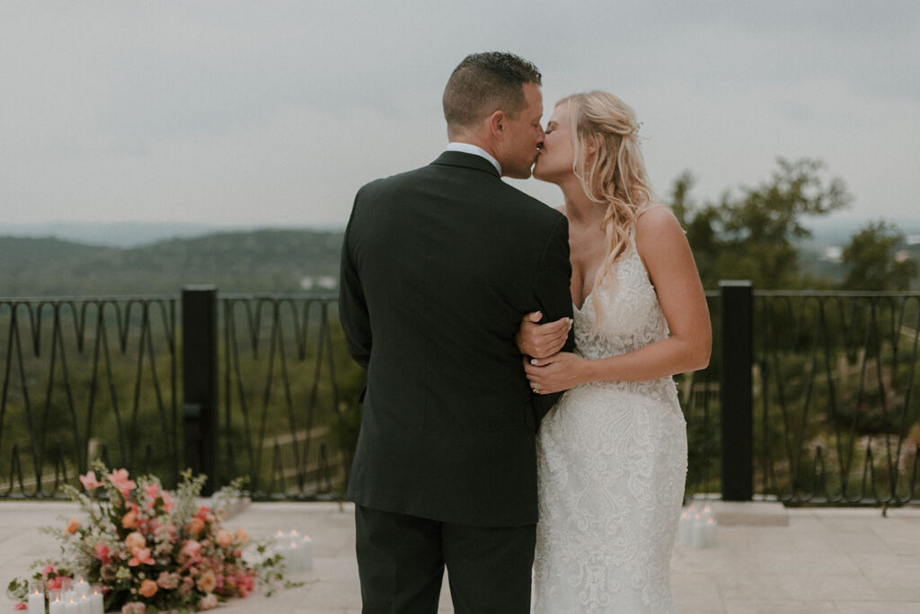 Bride and groom share a kiss outdoors on a terrace, framed by floral arrangements and candles, with Texas Hill Country views in the background.