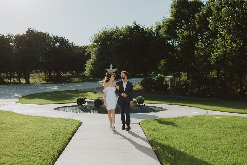 ChatGPT said:
Couple walking arm in arm down a sunlit pathway surrounded by vibrant green lawns, with a circular fountain and manicured landscaping in the background.