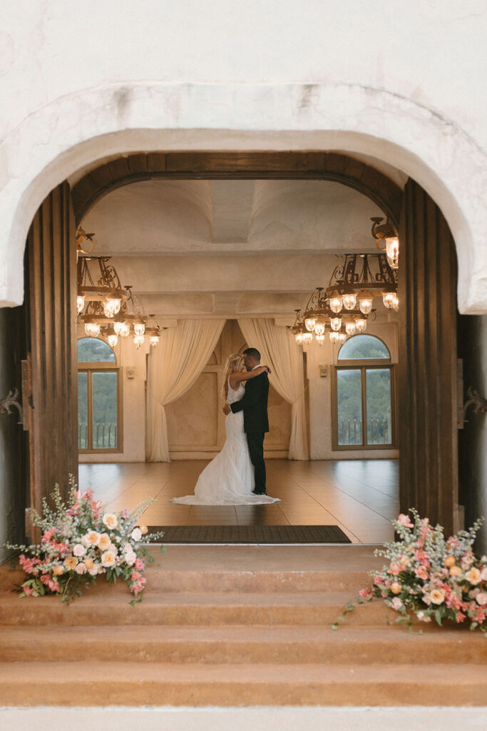 Bride and groom embrace in the center of a grand reception hall, framed by arched windows, draped curtains, and ornate chandeliers, with floral arrangements at the entrance.
