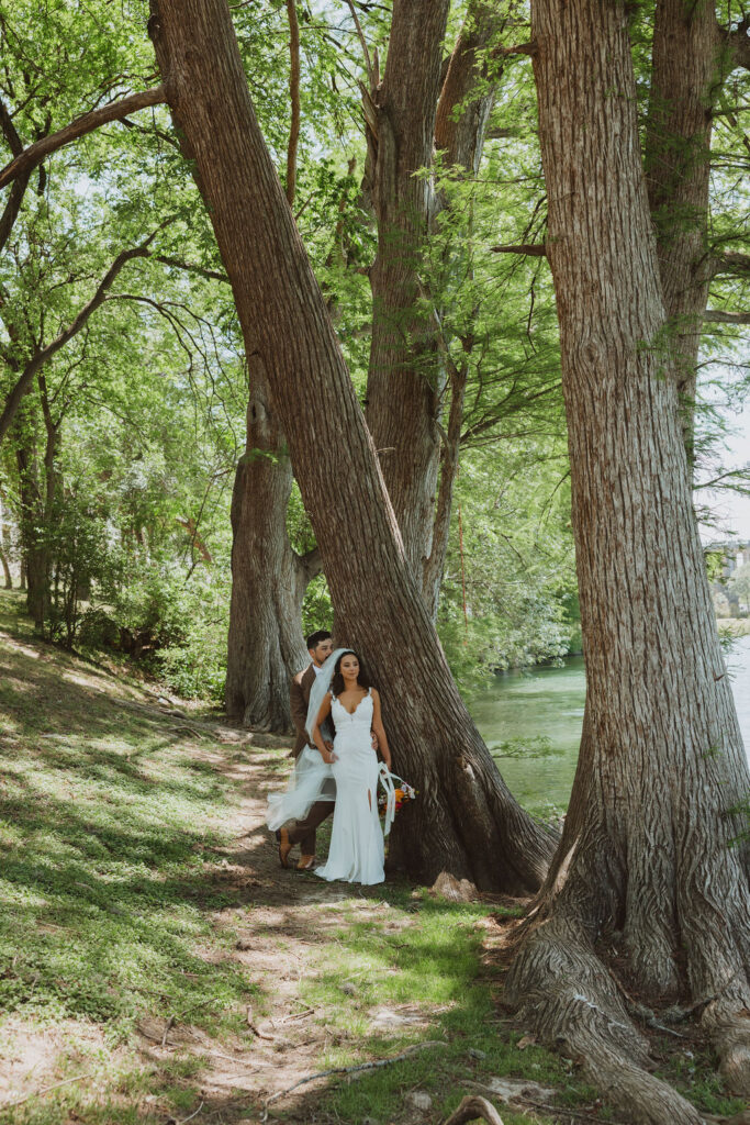 Bride and groom standing close together among tall trees along a calm riverbank, bride holding bouquet.
