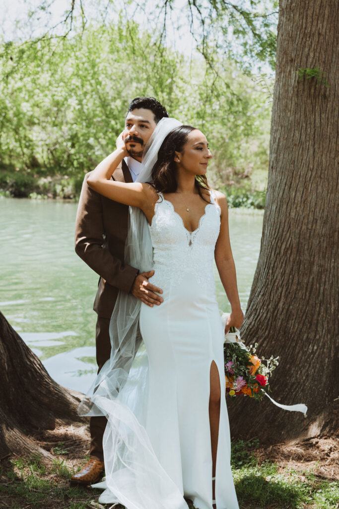 Bride holding bouquet and resting her hand on groom’s face while standing beside a tree by the river.