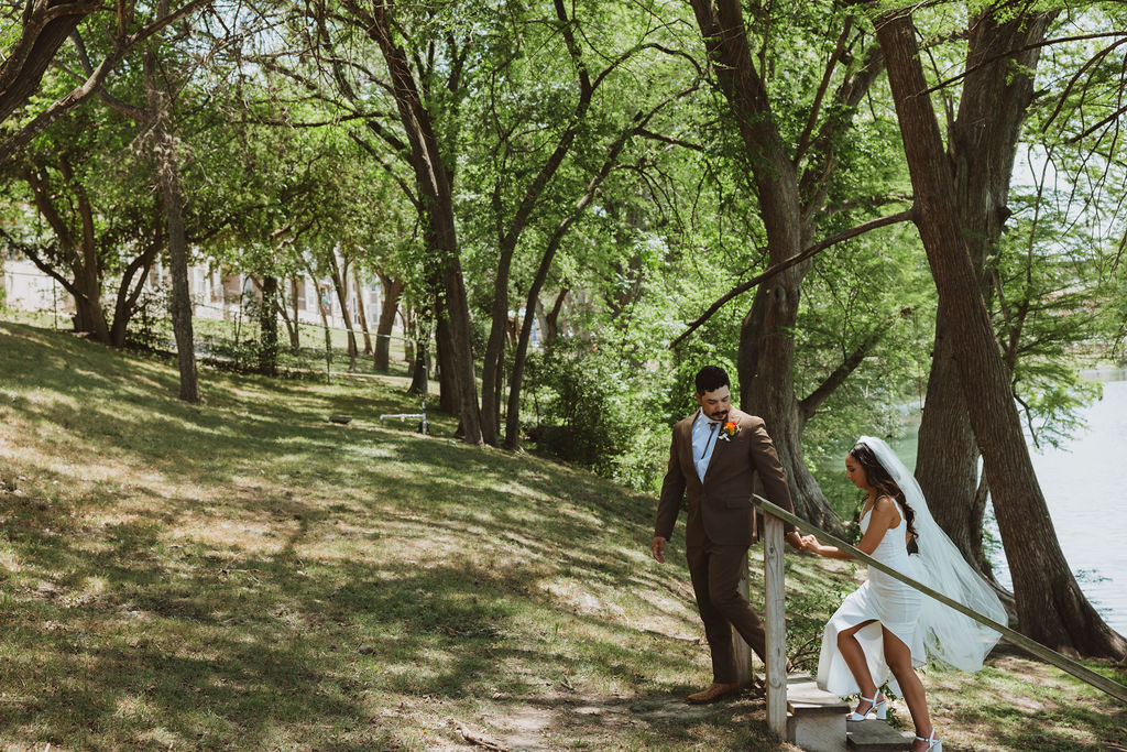 Groom helping bride up a small wooden staircase in a shaded riverside park.