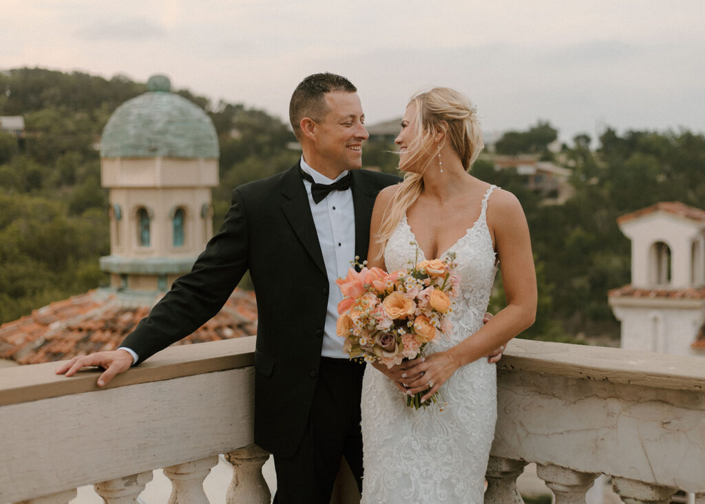 Bride and groom standing together on a balcony, smiling at each other, with terracotta rooftops and a green-domed tower in the scenic background.