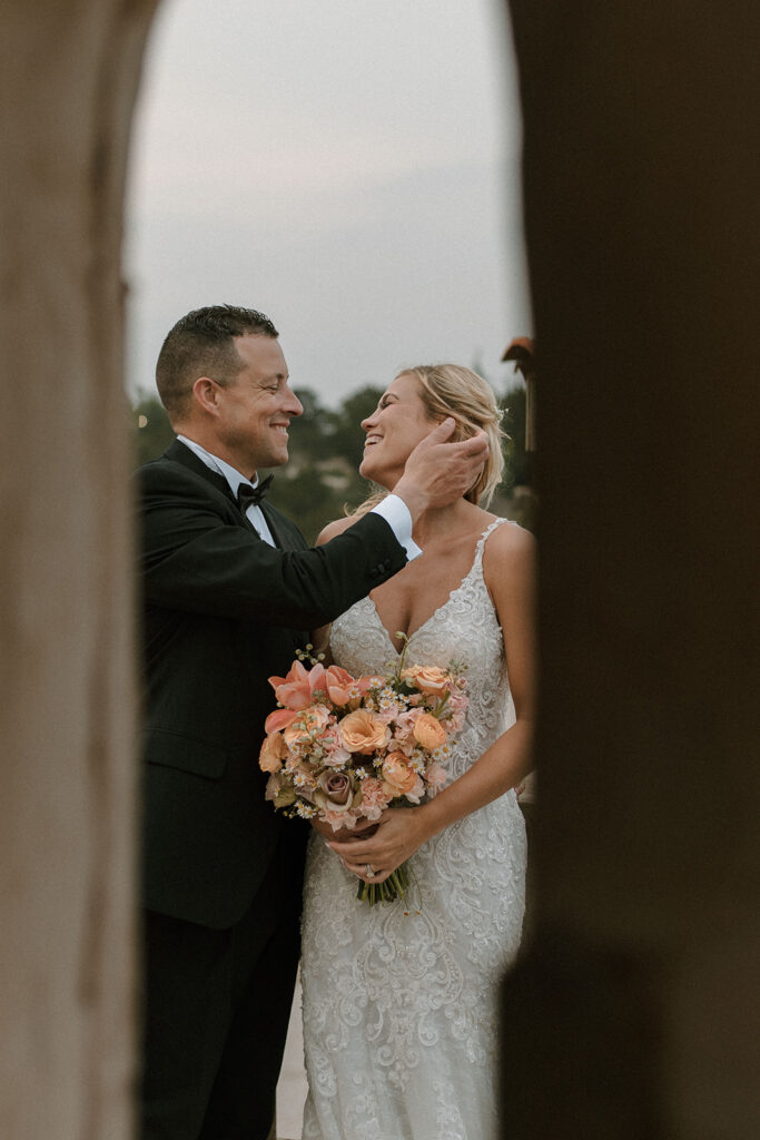 Groom smiling at bride while gently holding her face, both framed through an arched stone opening, with the bride holding a peach and blush bouquet.