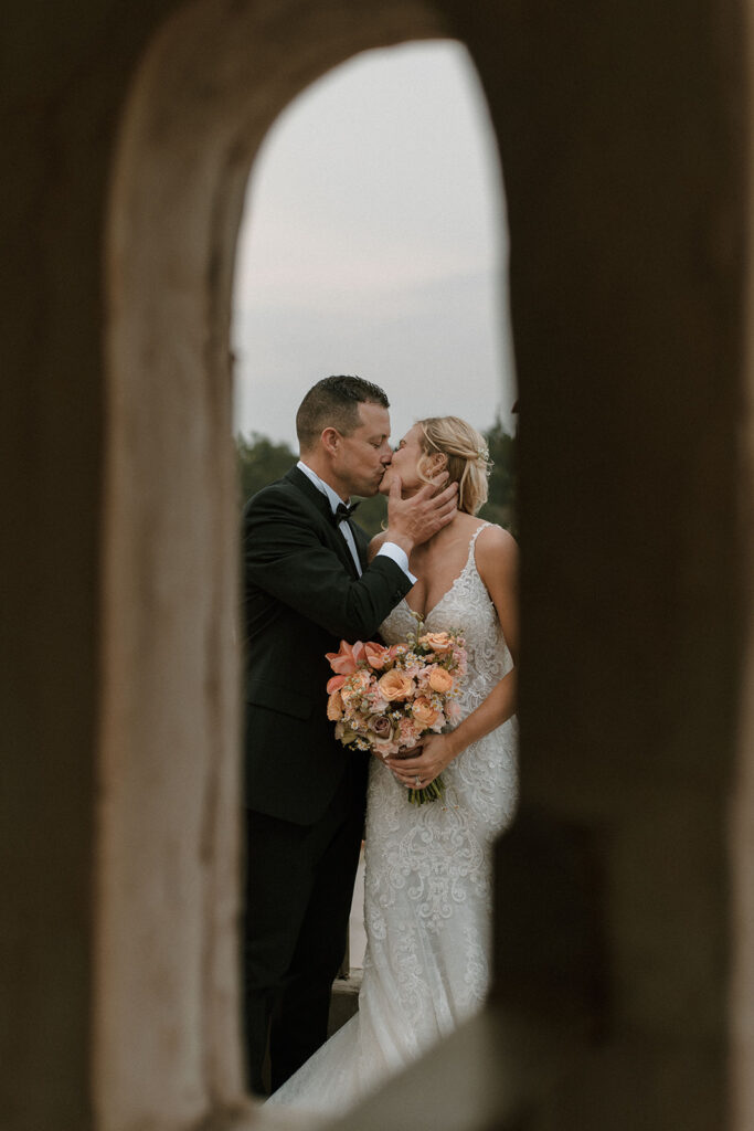 Romantic shot of bride and groom sharing a kiss, framed through an arched stone opening, with the bride holding a bouquet of peach and blush flowers.
