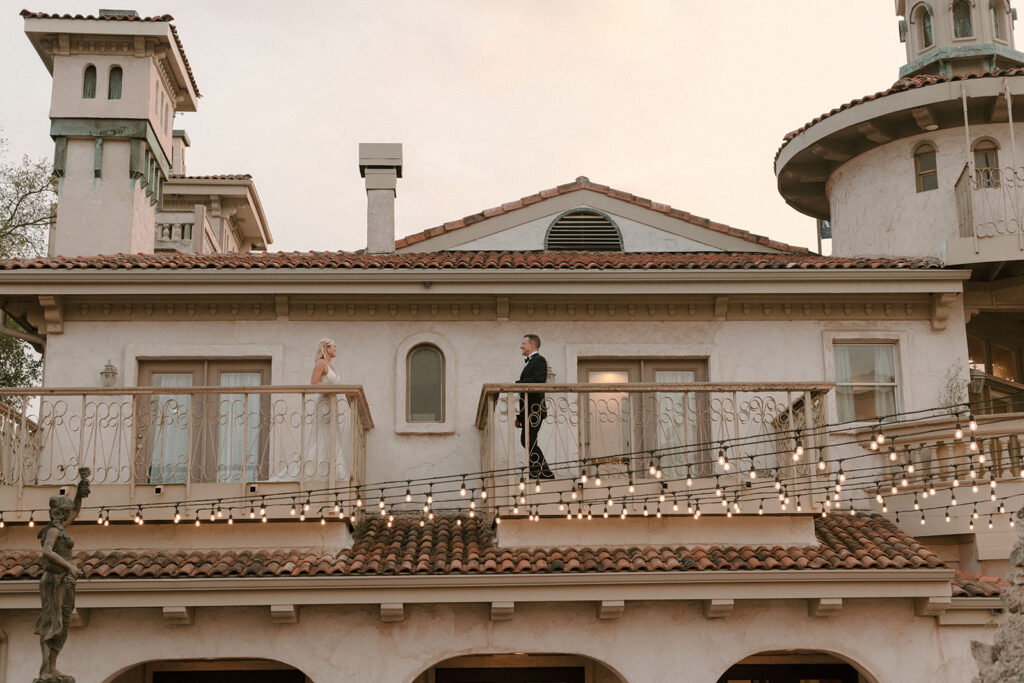 Wide shot of bride and groom standing on separate balconies of a Mediterranean-style villa at sunset, with strings of glowing café lights connecting the space between them.