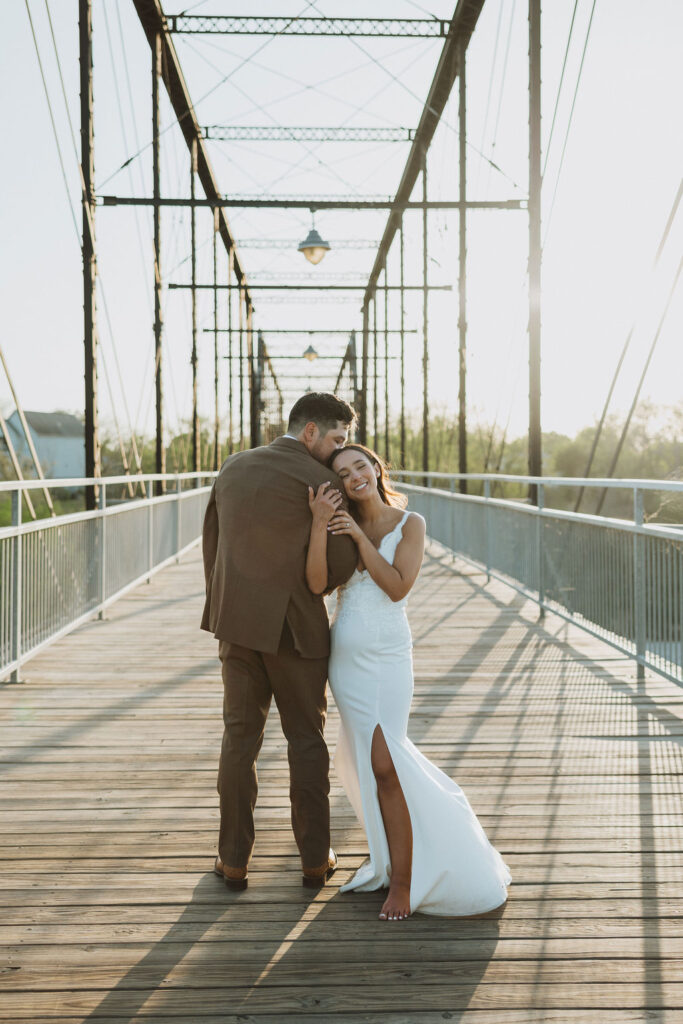 Groom embracing bride from behind while she smiles with eyes closed on a wooden bridge.