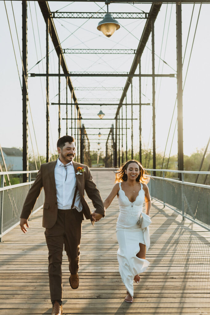 Bride and groom running hand in hand across a metal truss bridge at sunset.  