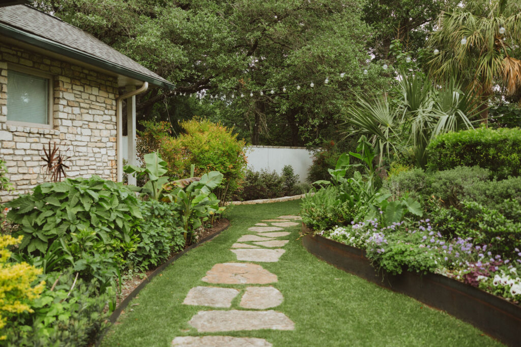 Stone pathway winding through a landscaped garden beside a stone building, lined with lush plants and flowers.
