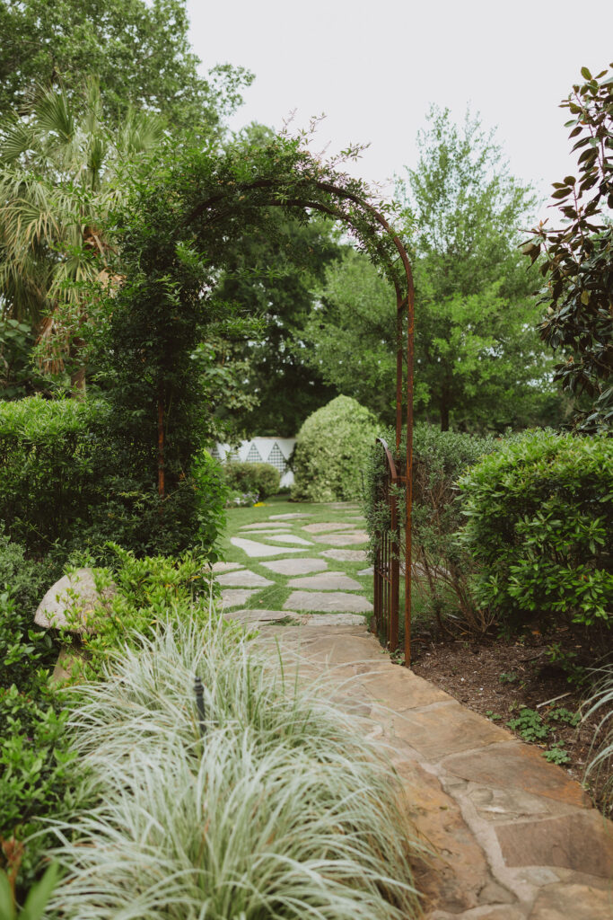  Stone pathway leading through a garden under a rustic metal arch covered in greenery.

