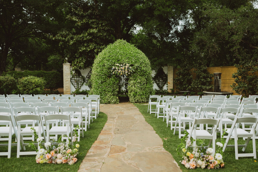 Outdoor wedding ceremony setup with rows of white chairs facing a large floral arch made of greenery and pastel flowers.
