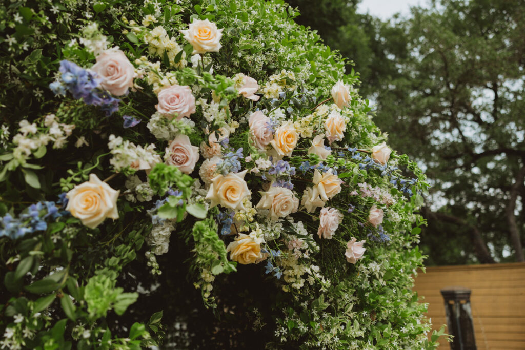 Close-up of a lush floral wedding arch decorated with pastel roses, blue delphiniums, and white blossoms surrounded by greenery.
