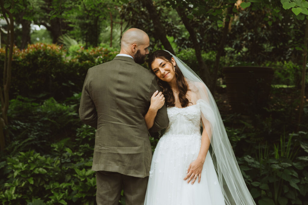  Bride resting her head on the groom’s shoulder, smiling softly while standing together in a shaded garden.