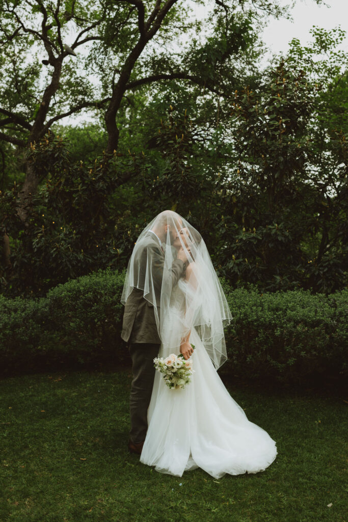Bride and groom kissing beneath the bride’s veil, with the bride holding a bouquet and surrounded by green foliage.