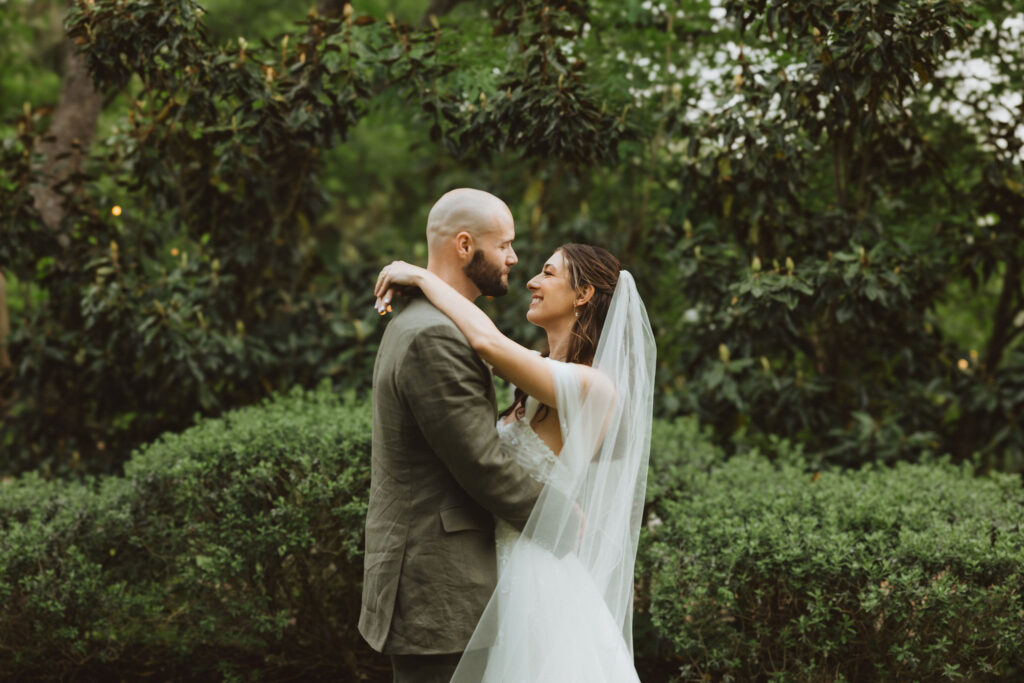 Bride and groom embracing and smiling at each other, standing in front of manicured hedges and greenery.