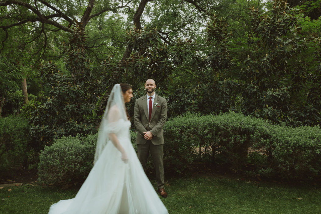 Groom standing with hands clasped, smiling as the bride walks toward him during their first look in a garden.