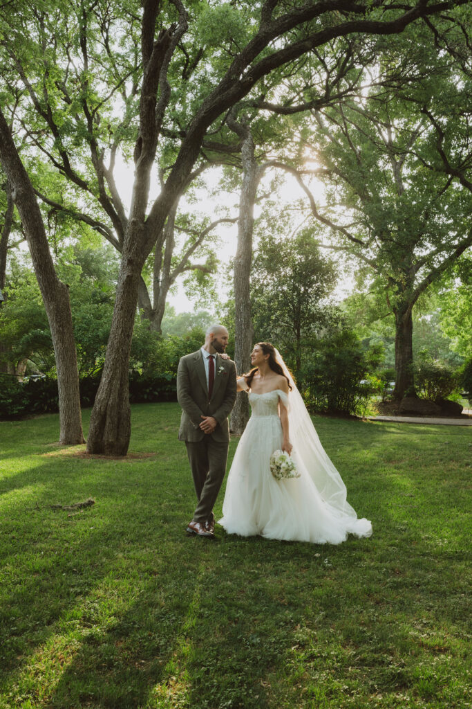 Bride and groom walking hand in hand on a green lawn, smiling at each other under tall trees with sunlight filtering through.