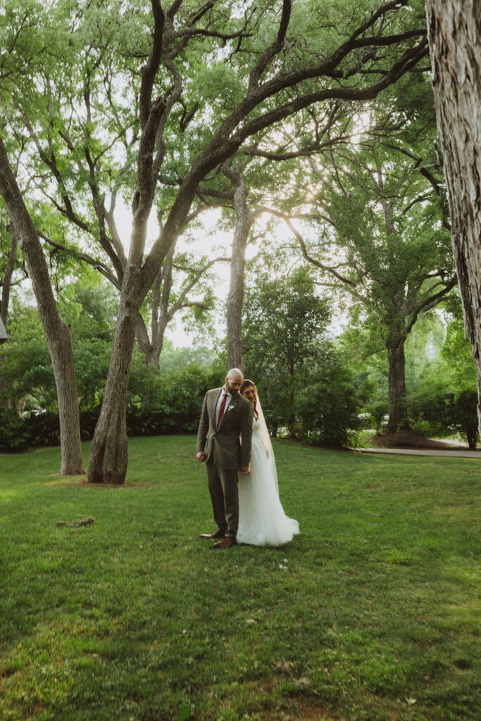Bride and groom standing close together in a lush green park, surrounded by tall trees with sunlight streaming through the branches.