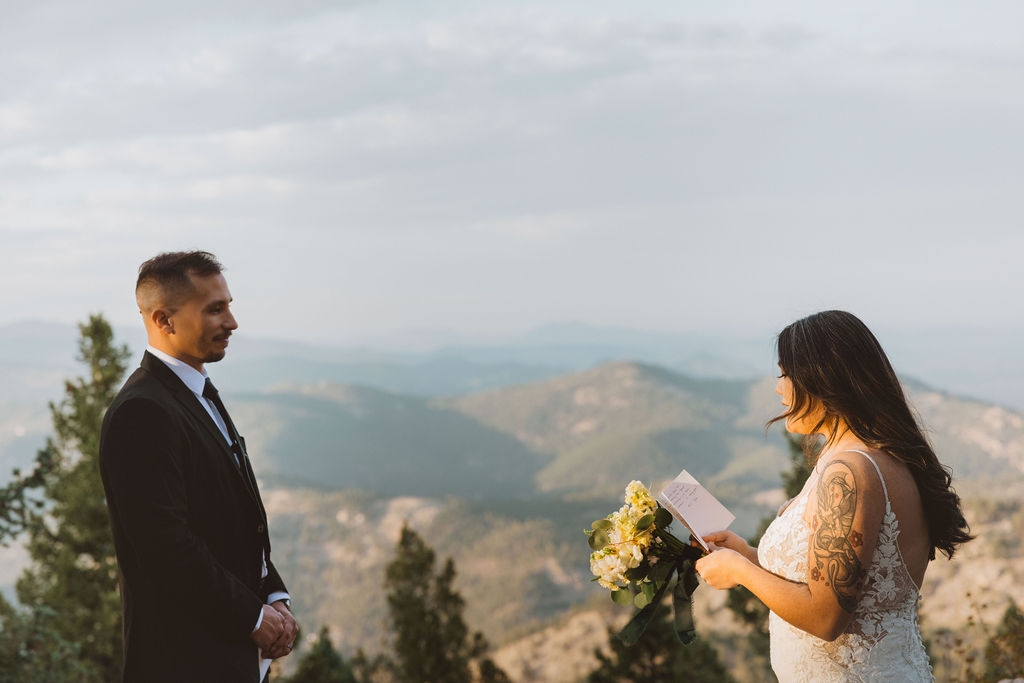A bride and groom exchange vows with mountains in the background as the sun is setting.