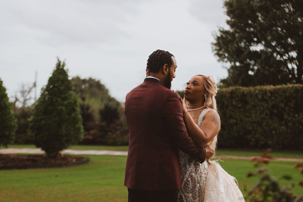 Bride and groom standing close, gazing into each other’s eyes in an open garden with dramatic, cloudy skies overhead.