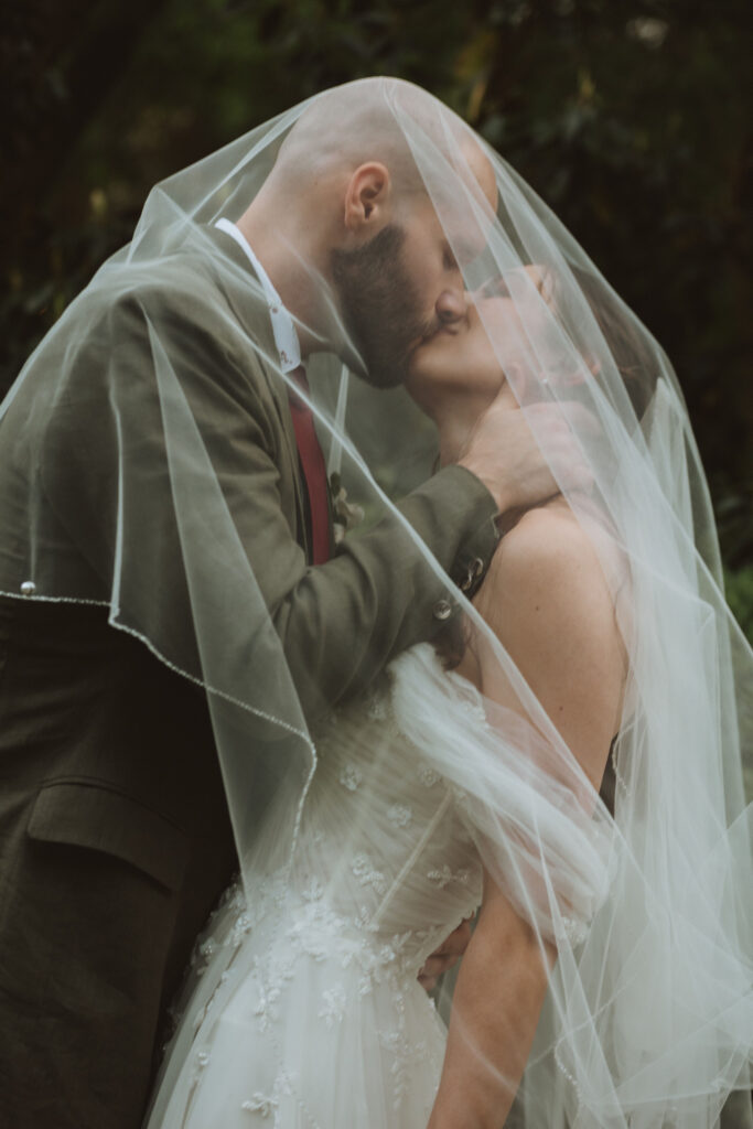 Bride and groom sharing a romantic kiss under the bride’s veil, with lush greenery in the background.