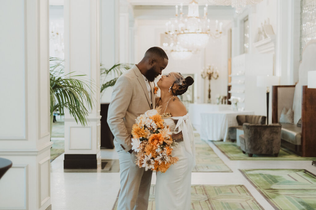 Groom leans in toward the bride holding a cascading bouquet of orange, peach, and white flowers in a luxurious hotel lobby.