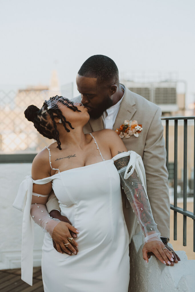 Groom kisses bride on the forehead while holding her close on a rooftop, the bride’s sheer pearl-detailed sleeves and tattoo visible.