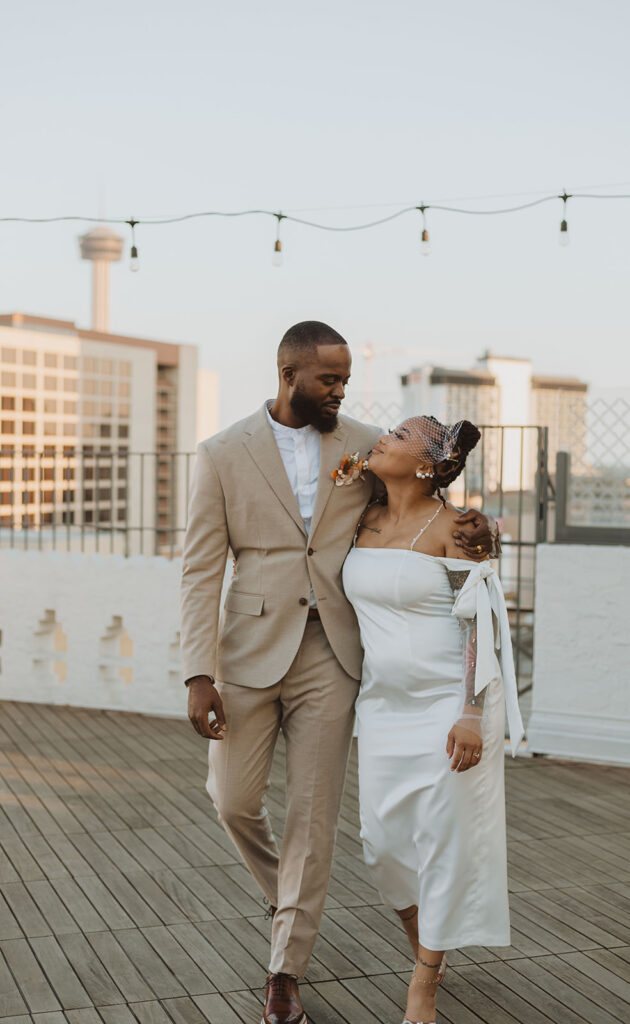 Groom in a tan suit and bride in an off-the-shoulder white dress with sheer, pearl-detailed sleeves walk arm-in-arm on a rooftop deck in San Antonio, with the Tower of the Americas visible in the background.