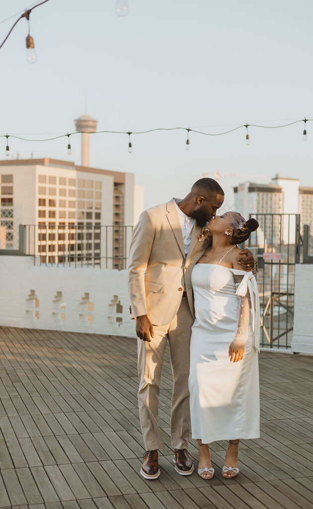 Groom and bride share a kiss on a rooftop with city buildings and the Tower of the Americas in the background.