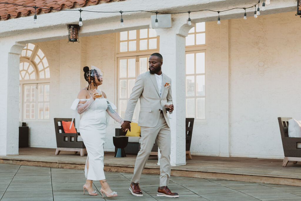 Groom in a tan suit and bride in an off-the-shoulder white dress with sheer, pearl-detailed sleeves walk on a rooftop deck in San Antonio, 