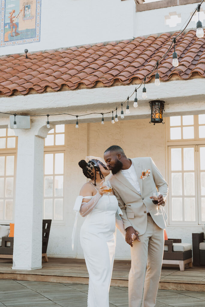 Bride and groom kiss while holding drinks on a rooftop patio with string lights and Spanish tile roofing above them.
