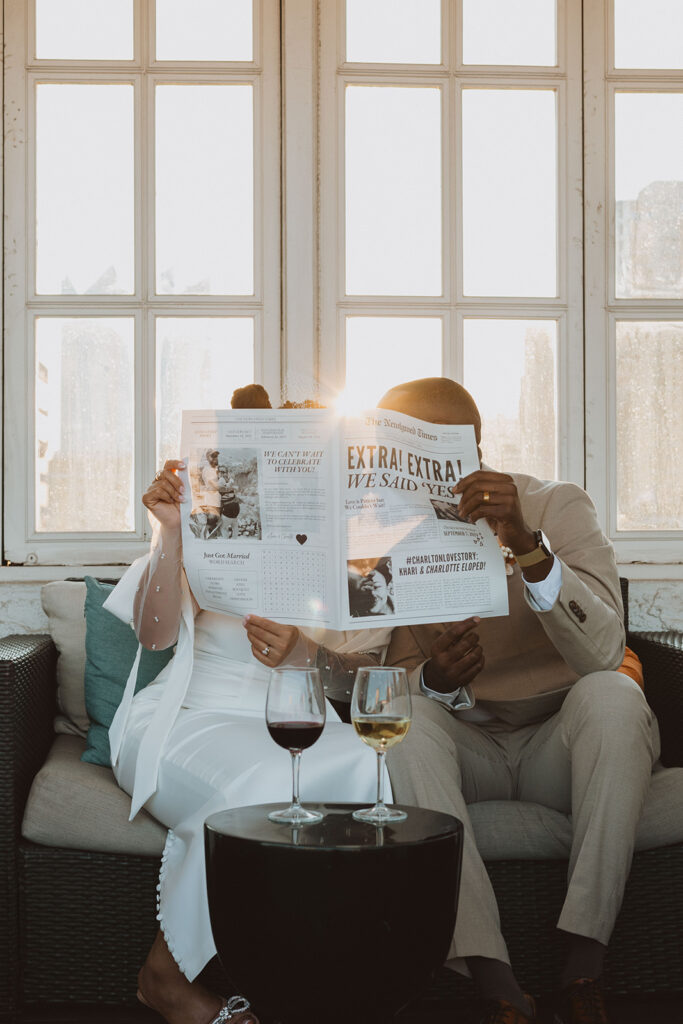 Bride and groom sit together on a rooftop lounge, hiding their faces behind a custom newspaper announcing their elopement, with wine glasses on a table in front of them.