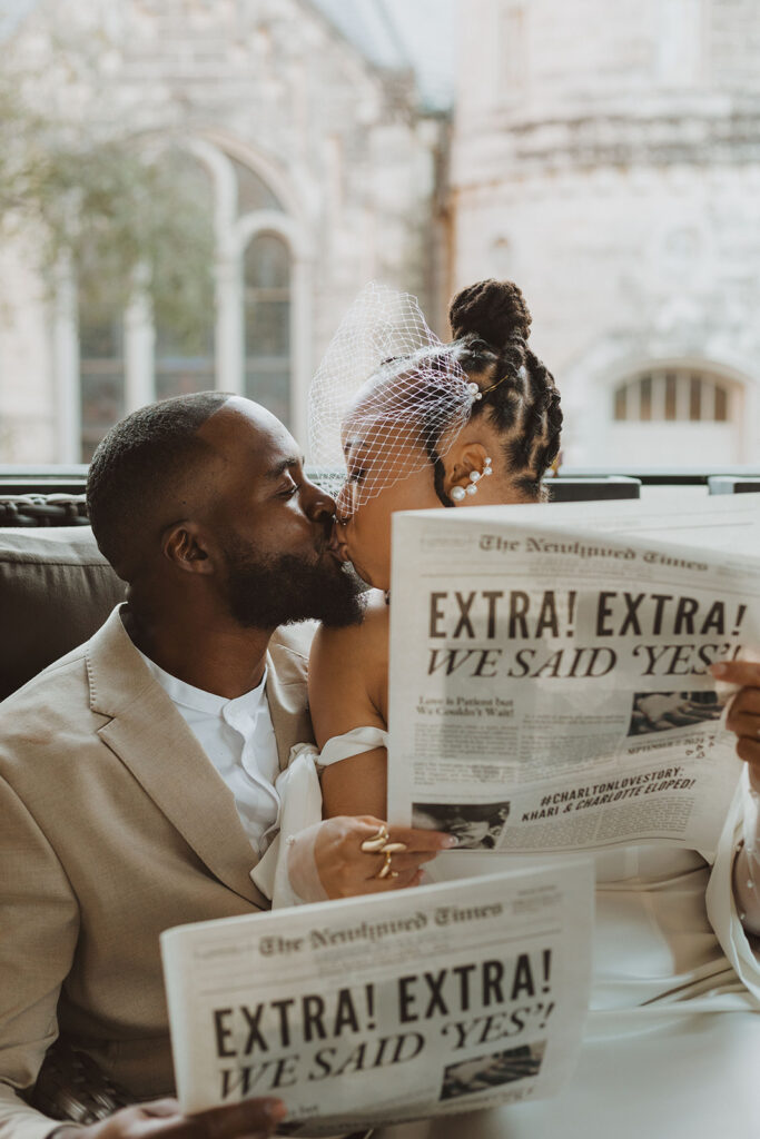 Bride and groom kiss while holding newspapers with a headline announcing “We Said Yes,” seated in front of a window with a historic building in the background.