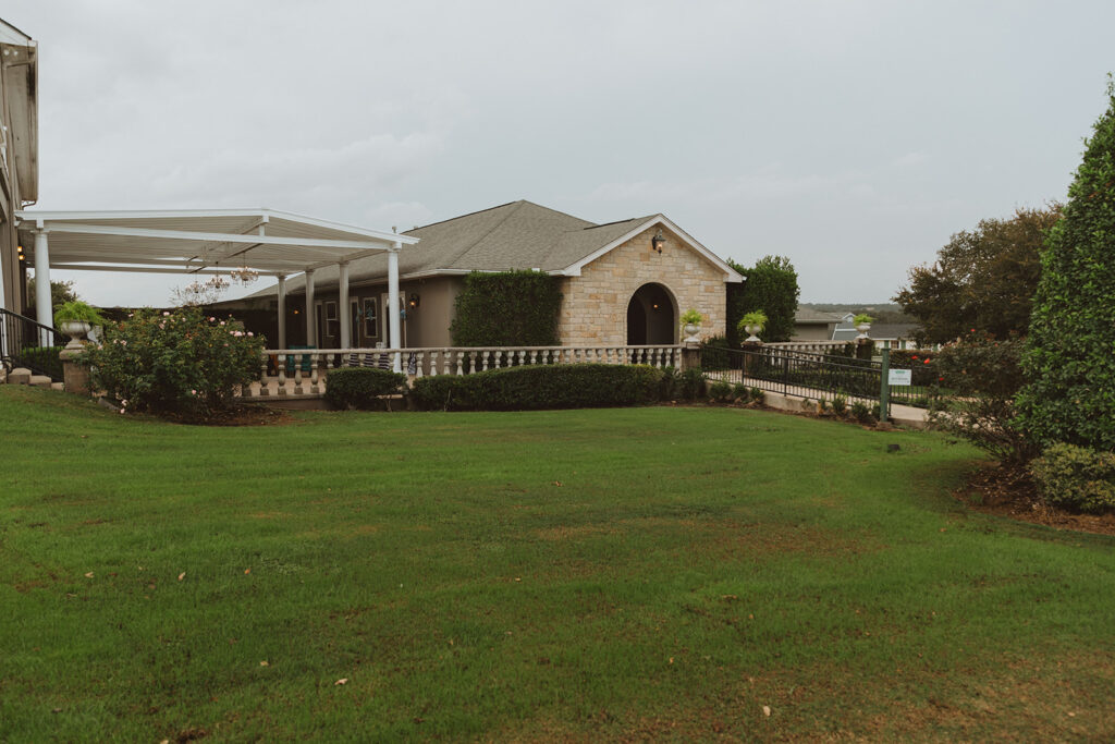 Side view of a wedding venue building with a covered patio, stone accents, manicured landscaping, and open grassy space in the foreground.  