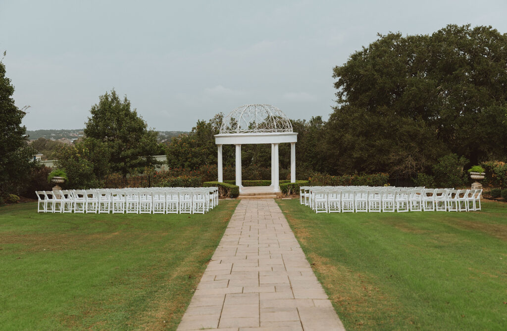 Outdoor wedding ceremony setup with rows of white chairs facing a white columned gazebo, surrounded by lush green lawns and trees.