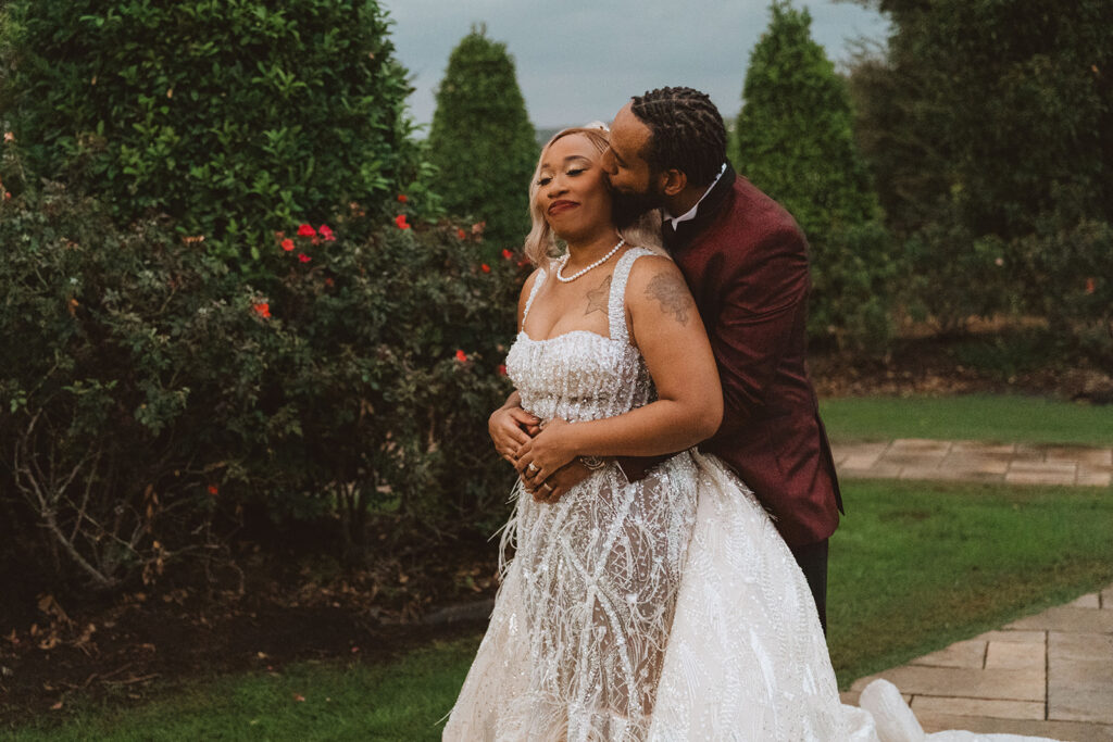 Groom embracing the bride from behind and kissing her cheek as she smiles softly, surrounded by manicured bushes and red flowers.  
